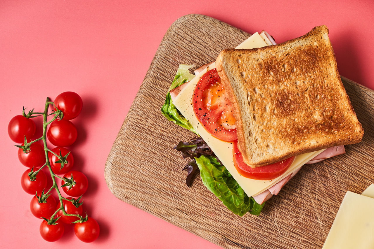 Toast-Sandwich mit Käse, Schinken, Tomate und Salat auf einem Holzbrett, daneben eine Rispe Kirschtomaten auf rosa Untergrund.