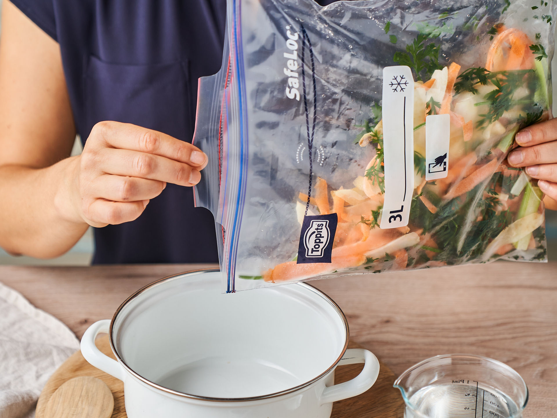 Eine Frau hält einen Toppits SafeLoc Gefrierbeutel mit Gemüseschalen und Kräutern über einen leeren weißen Kochtopf. Auf dem Tisch liegen ein Holzlöffel, ein Messbecher mit Wasser, Lorbeerblätter und Wacholderbeeren in kleinen Schalen