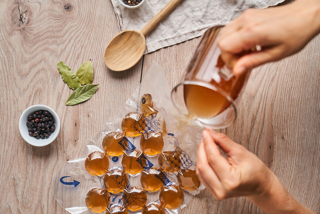 Eine Person füllt  Brühe, aus einem Glaskanne in eine Toppits Eiskugel Beutel mit mehreren Kammern. Auf dem Holztisch liegen Lorbeerblätter, eine Schale mit Wacholderbeeren, ein Holzlöffel und Pfefferkörner