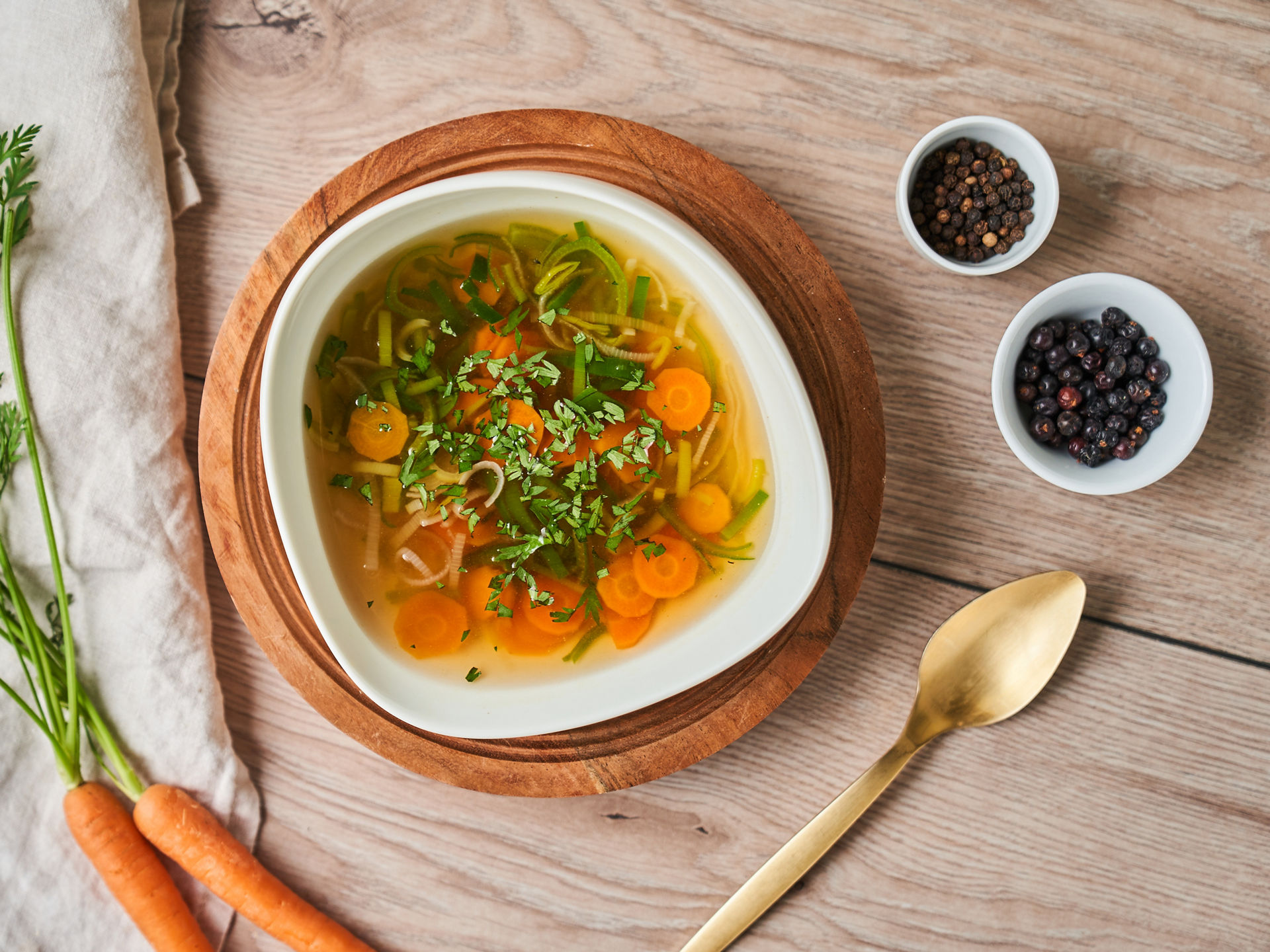 Eine Schale Gemüsesuppe mit Karottenscheiben, Lauch und frischer Petersilie steht auf einem Holzbrett. Daneben liegen frische Karotten, ein goldener Löffel und kleine Schalen mit Pfefferkörnern und Wacholderbeeren auf einem hellen Holztisch. 