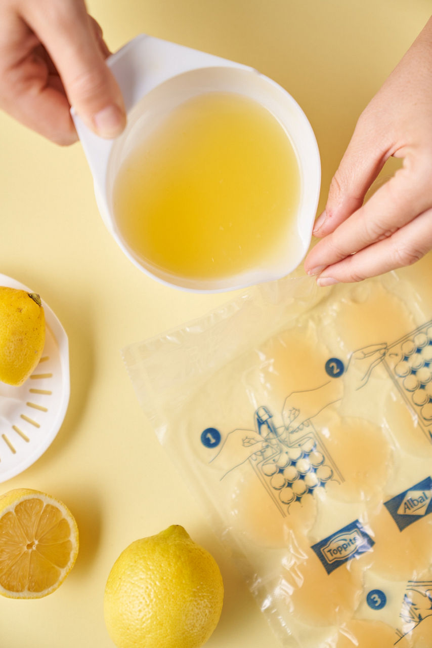 Eine Person gießt frisch gepressten Zitronensaft aus einem weißen Messbecher in einen Toppits Eiskugelbeutel. Auf dem gelben Untergrund liegen außerdem eine Zitronenpresse, eine halbe und eine ganze Zitrone