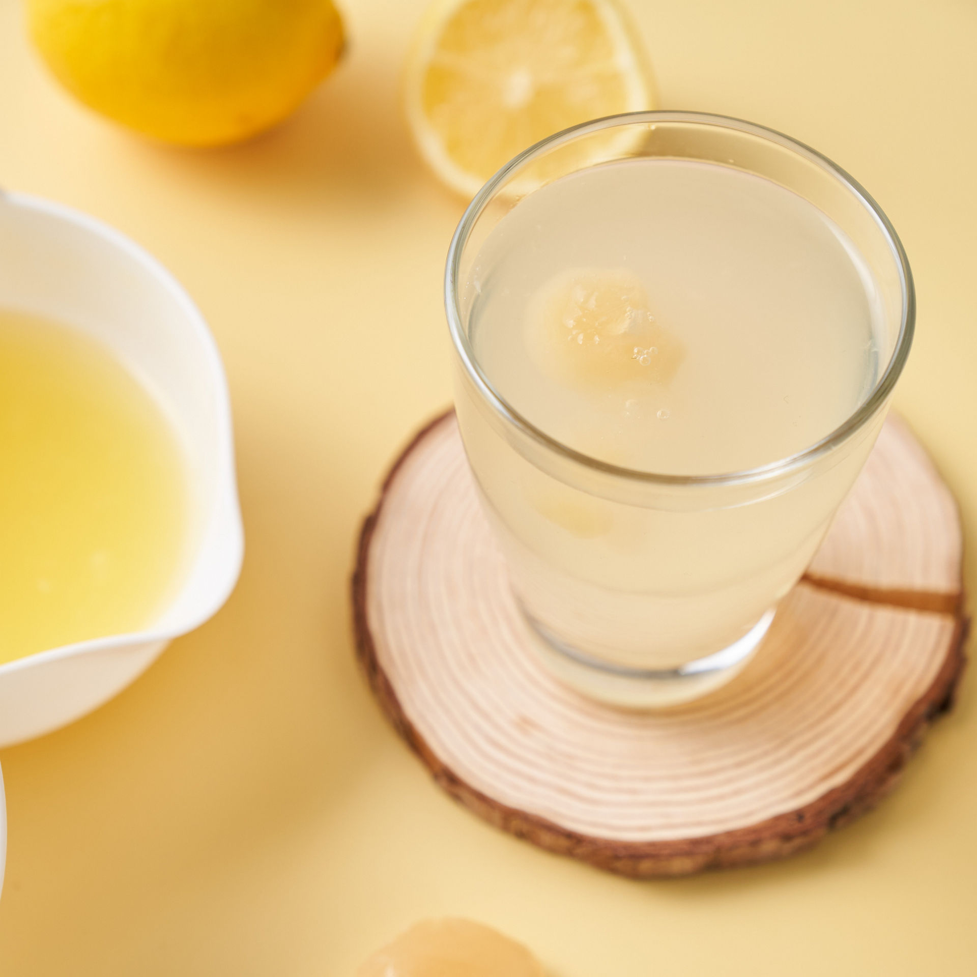Ein Glas mit Wasser und Zitroneneiskugeln steht auf einer Holzunterlage. Daneben liegen zwei Zitroneneiskugeln, ein weißer Messbecher mit Zitronensaft sowie eine ganze und eine halbe Zitrone auf gelbem Untergrund.