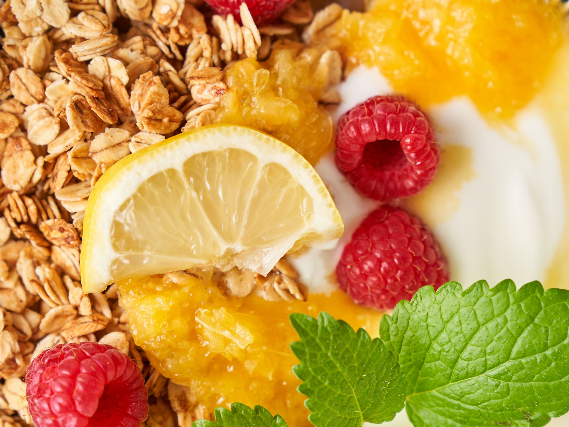 Nahaufnahme einer Schale mit Joghurt, Granola, Himbeeren, Zitronenscheiben, Orangenpüree und frischer Minze.