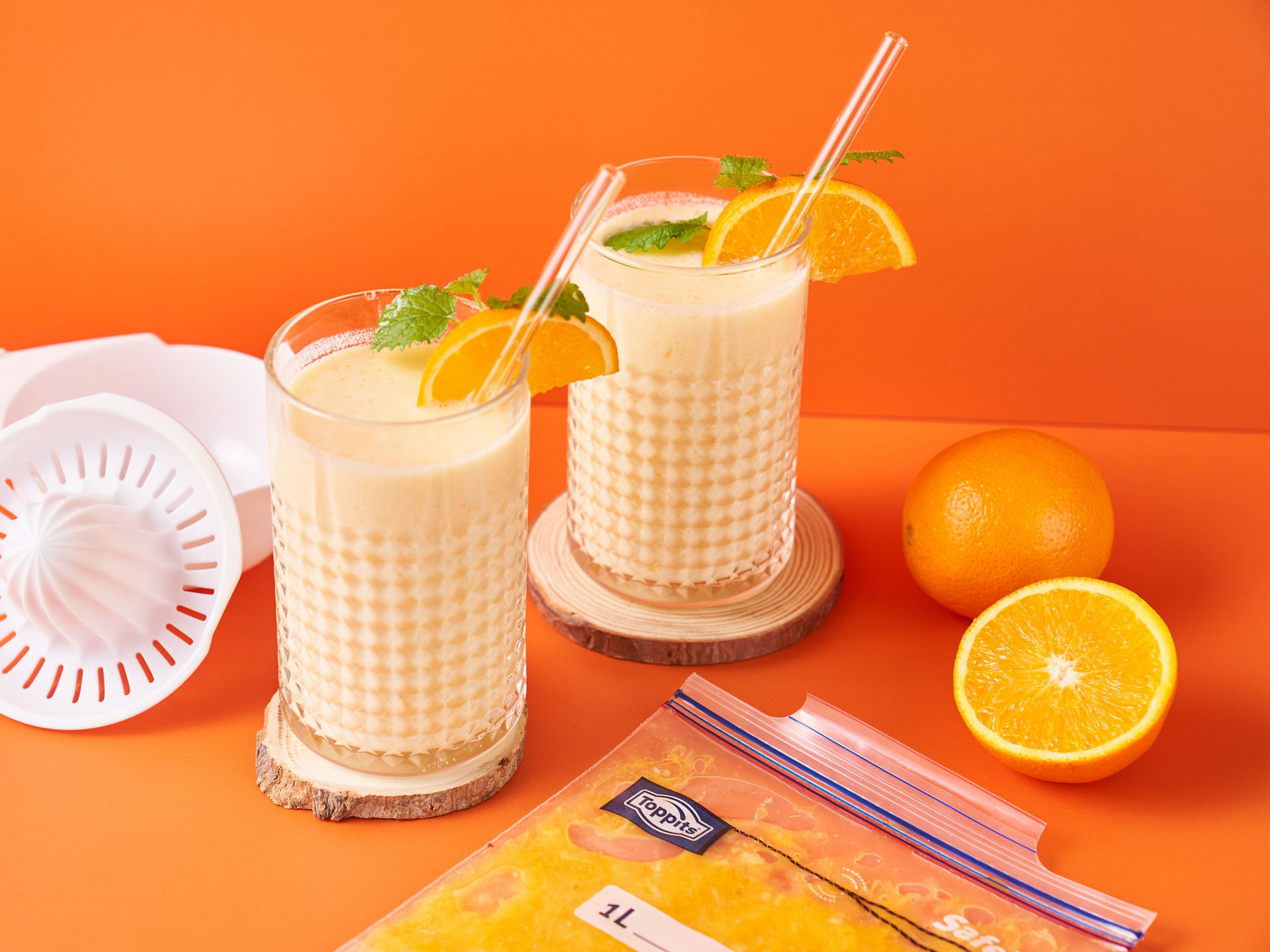 Zwei Gläser Orangen-Milchshake mit Orangenscheibe und Minze, daneben Orangen, ein Zitruspressaufsatz und ein Toppits SafeLoc Gefrierbeutel mit Orangenpüree 