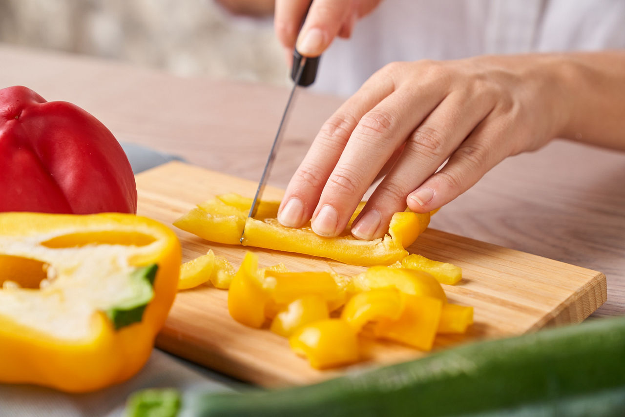 Eine Hand schneidet mit einem Messer gelbe Paprika in Streifen auf einem Holzbrett. Im Vordergrund liegen eine aufgeschnittene gelbe Paprika, eine rote Paprika und ein Stück Zucchini.