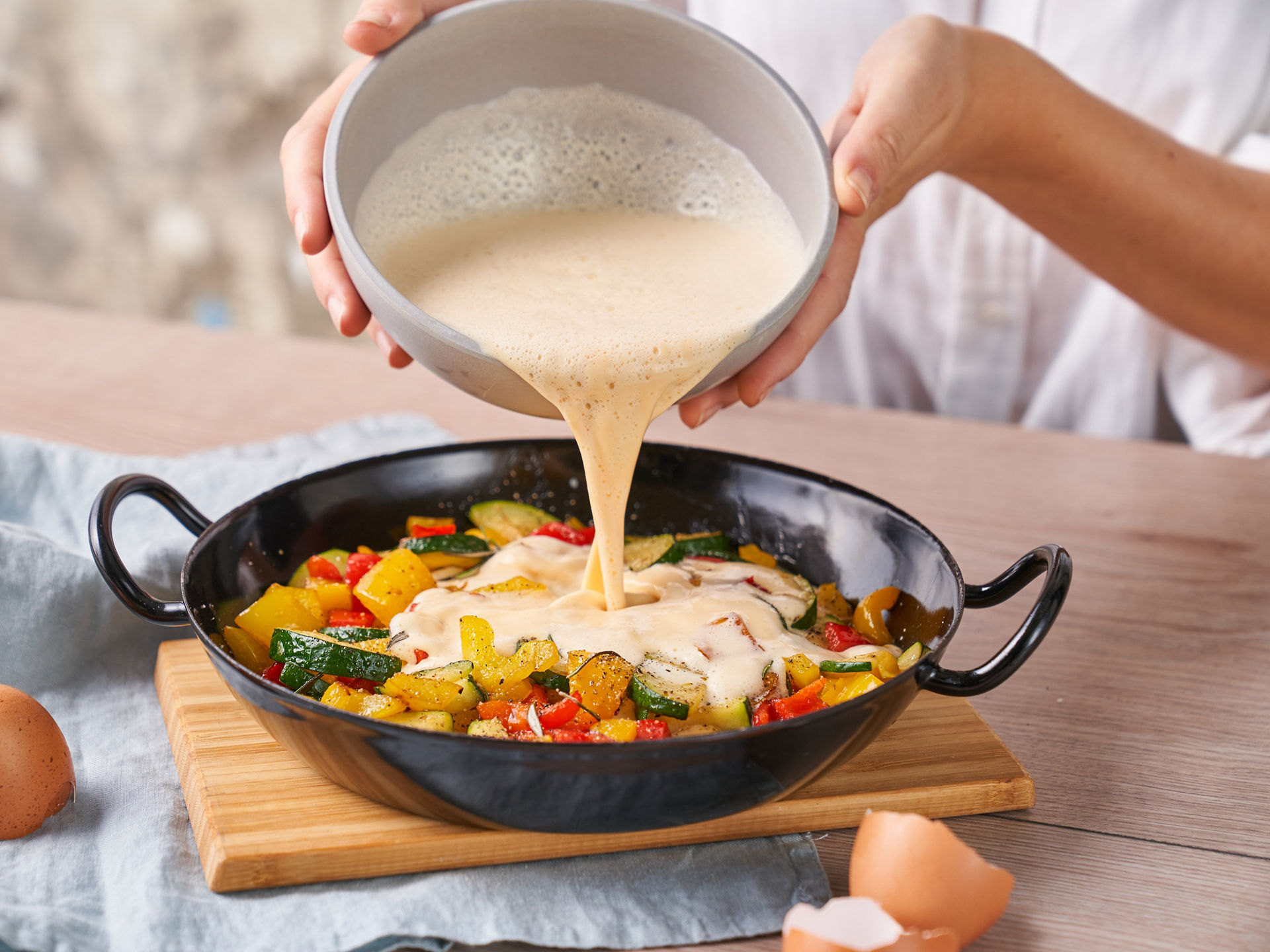Eine Person gießt eine schaumige Eiermischung aus einer Schüssel in eine schwarze Pfanne mit geschnittenem Gemüse wie Zucchini, gelber und roter Paprika. Auf dem Tisch liegen Eierschalen und ein Holzbrett