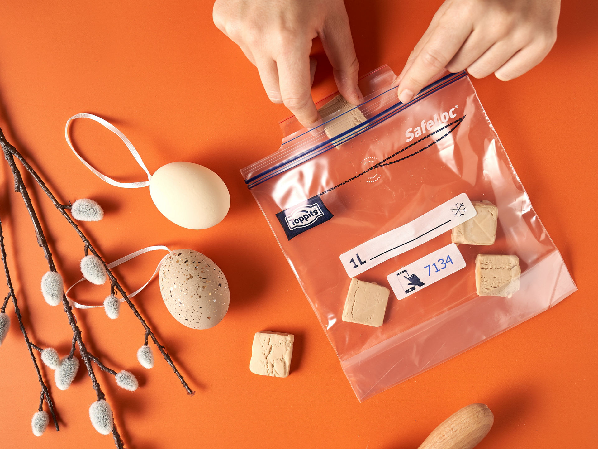 Zwei Hände verschließen einen Toppits SafeLoc Beutel mit mehreren Stücken Hefe auf einem orangefarbenen Untergrund. Neben dem Beutel liegen ein Weidenzweig mit Kätzchen, zwei Ostereier und ein Nudelholz 
