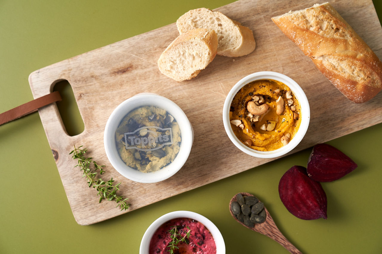 Drei kleine Schalen mit Hummus in verschiedenen Sorten stehen auf einem Holzbrett, dazu Baguette, frische Kräuter, Rote Bete und Kürbiskerne 