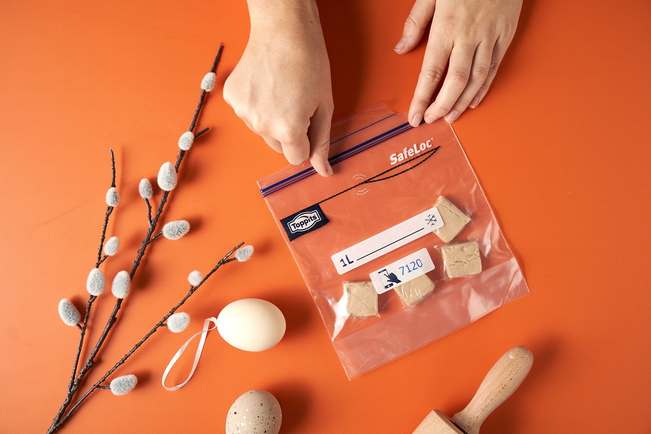 Zwei Hände verschließen einen Toppits SafeLoc Beutel mit mehreren Stücken frischer Hefe auf einem orangefarbenen Untergrund. Daneben liegen ein Weidenzweig mit Kätzchen, ein Osterei und ein Nudelholz