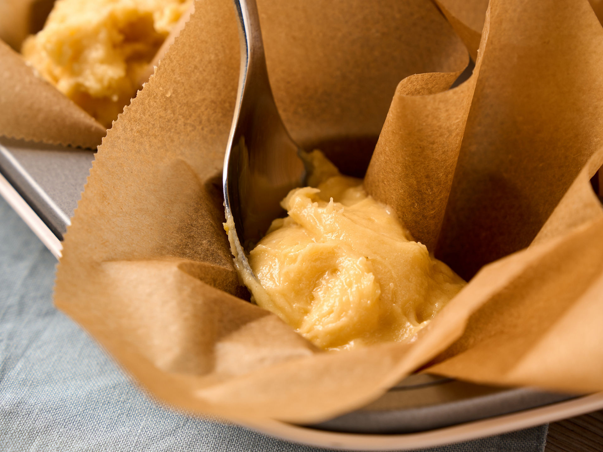 Ein Löffel füllt hellen Muffinteig in ein braunes Papierförmchen in einem Muffinblech