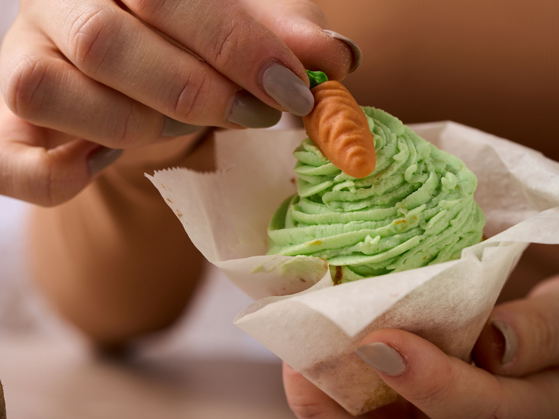 Nahaufnahme einer Hand, die eine kleine Marzipan-Möhre auf einen Muffin mit grünem Frosting setzt, Muffin in braunem Papierförmchen 
