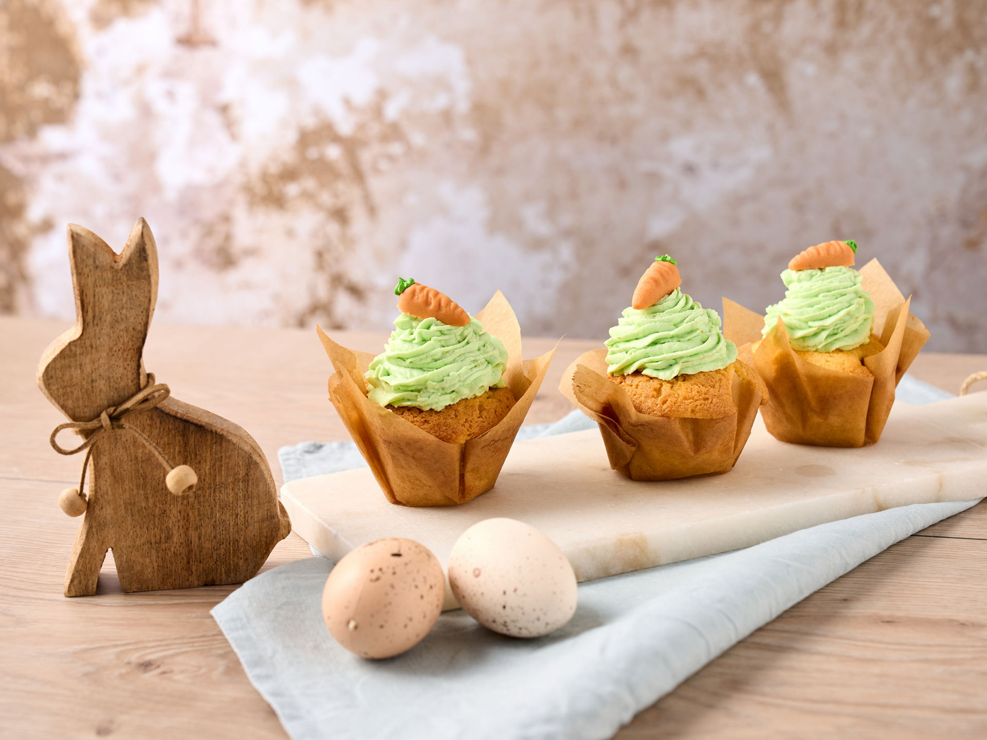 Drei Muffins mit grünem Frosting und Marzipan-Möhren als Dekoration, auf einer weißen Platte, daneben Deko-Ostereier und ein Holz-Osterhase 