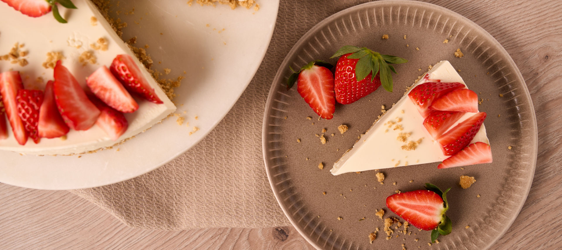 Ein Stück Käsekuchen mit Erdbeeren auf einem Teller, daneben die restliche Torte auf einer Tortenplatte. Beide stehen auf einem Holztisch