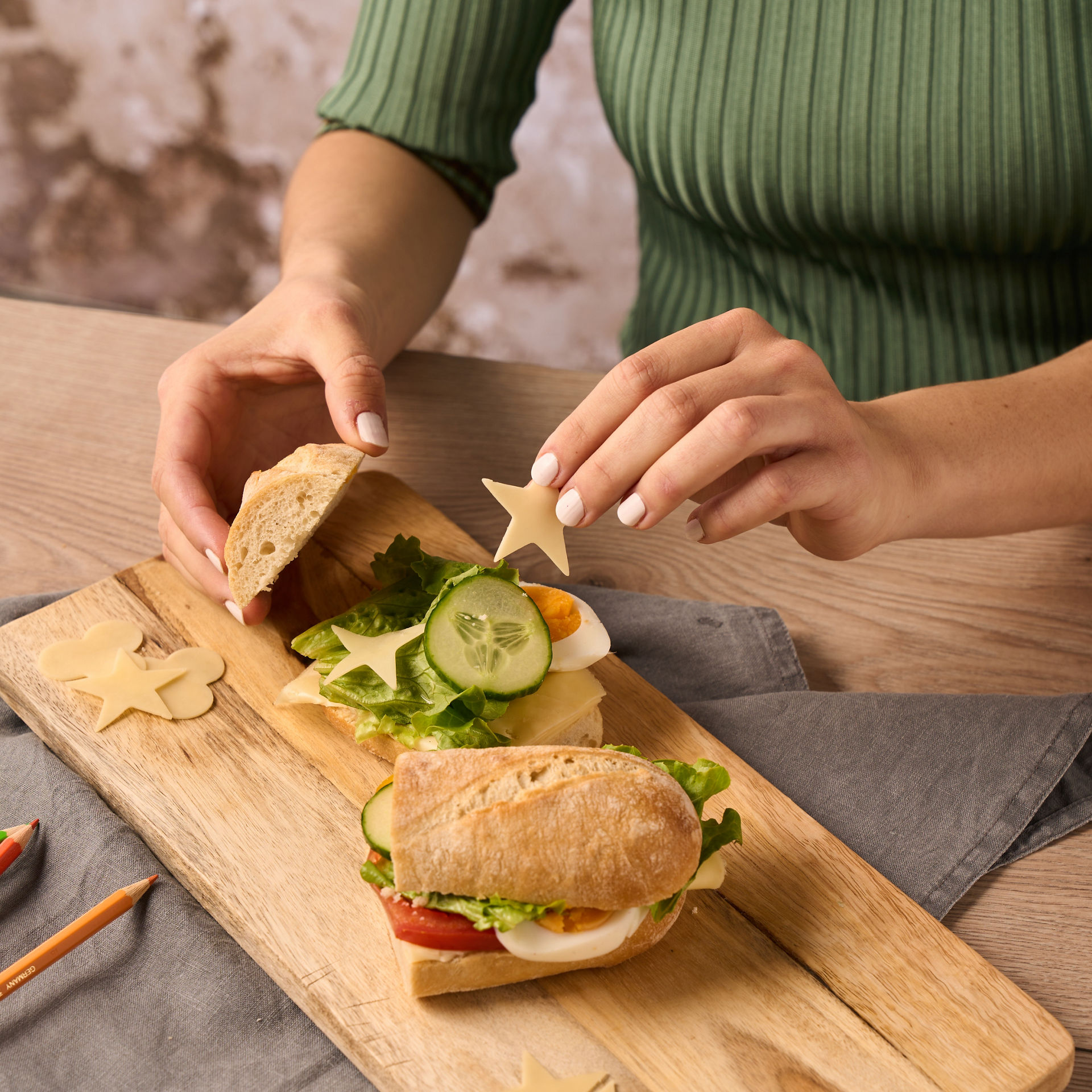 Eine Person in einem grünen Oberteil bereitet ein belegtes Brötchen auf einem Holzbrett zu.