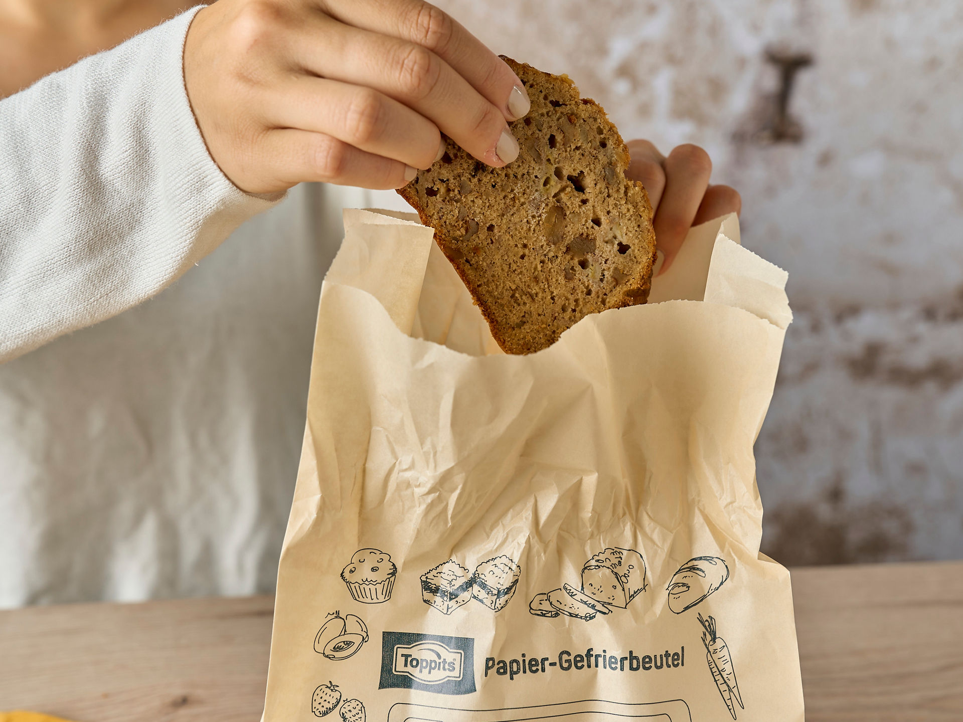 Eine Hand hält eine Scheibe Bananenbrot und legt sie in einen Toppits Papier-Gefrierbeutel