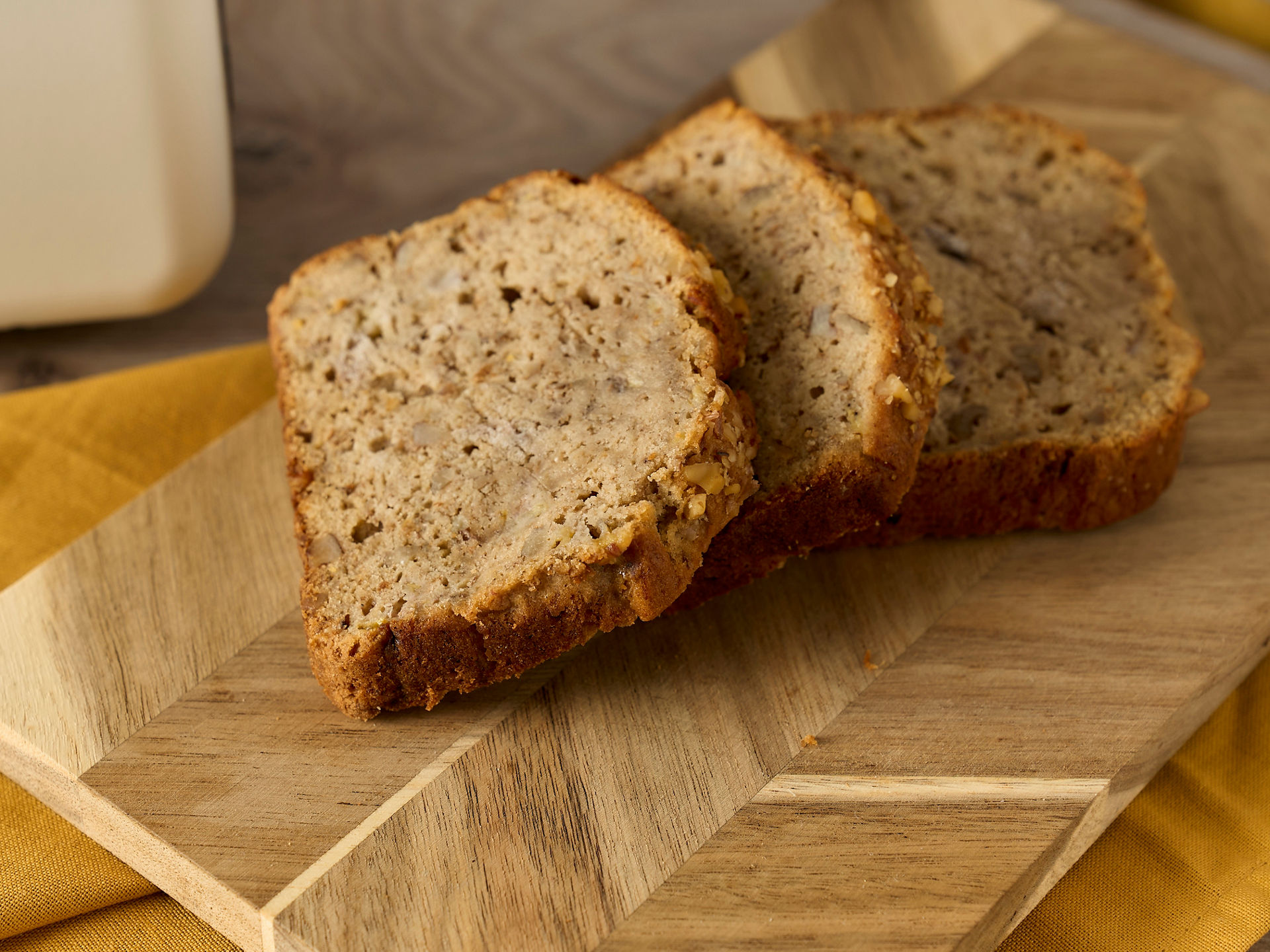 Drei Scheiben Bananenbrot liegen auf einem Holzbrett, das auf einem gelben Tuch platziert ist 