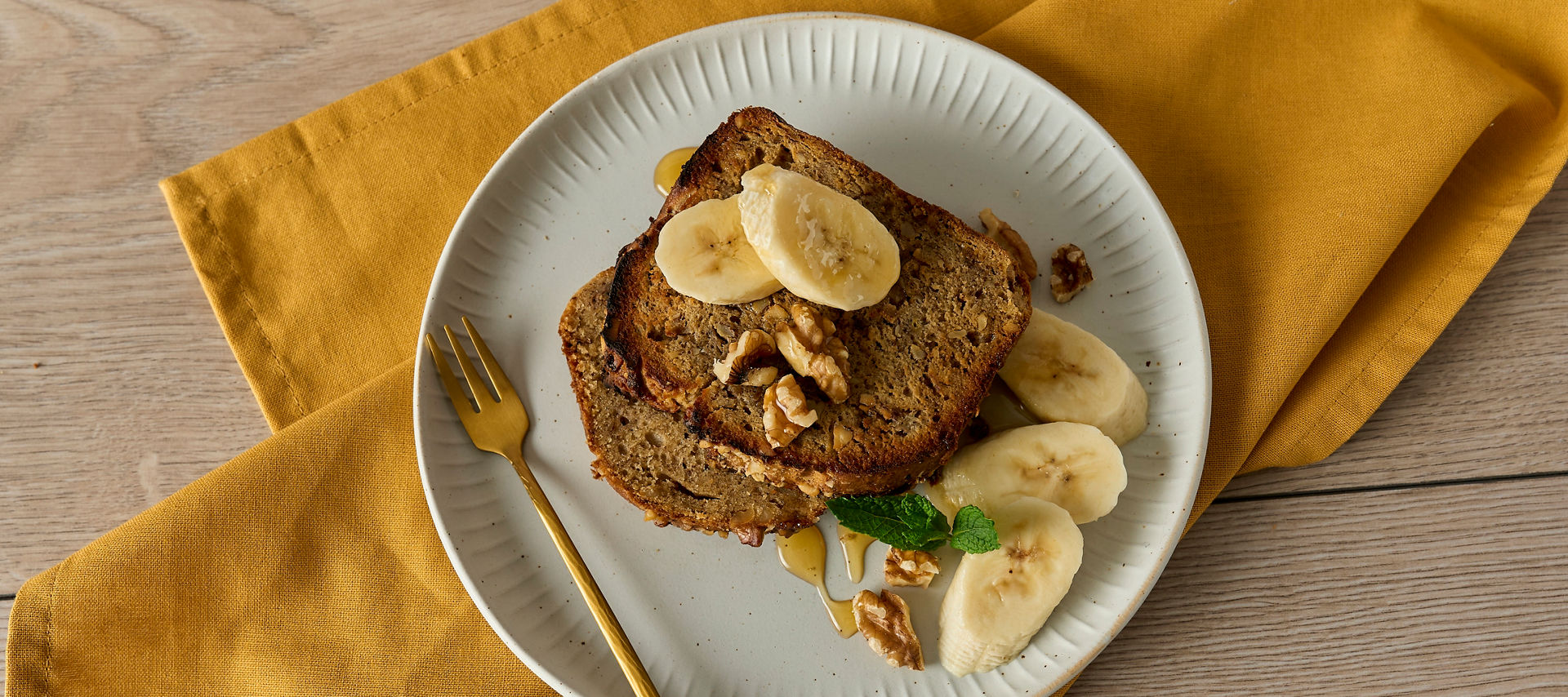 Zwei Scheiben Bananenbrot auf einem Teller, garniert mit Bananenscheiben, Walnüssen und Minze, auf einem gelben Tuch serviert