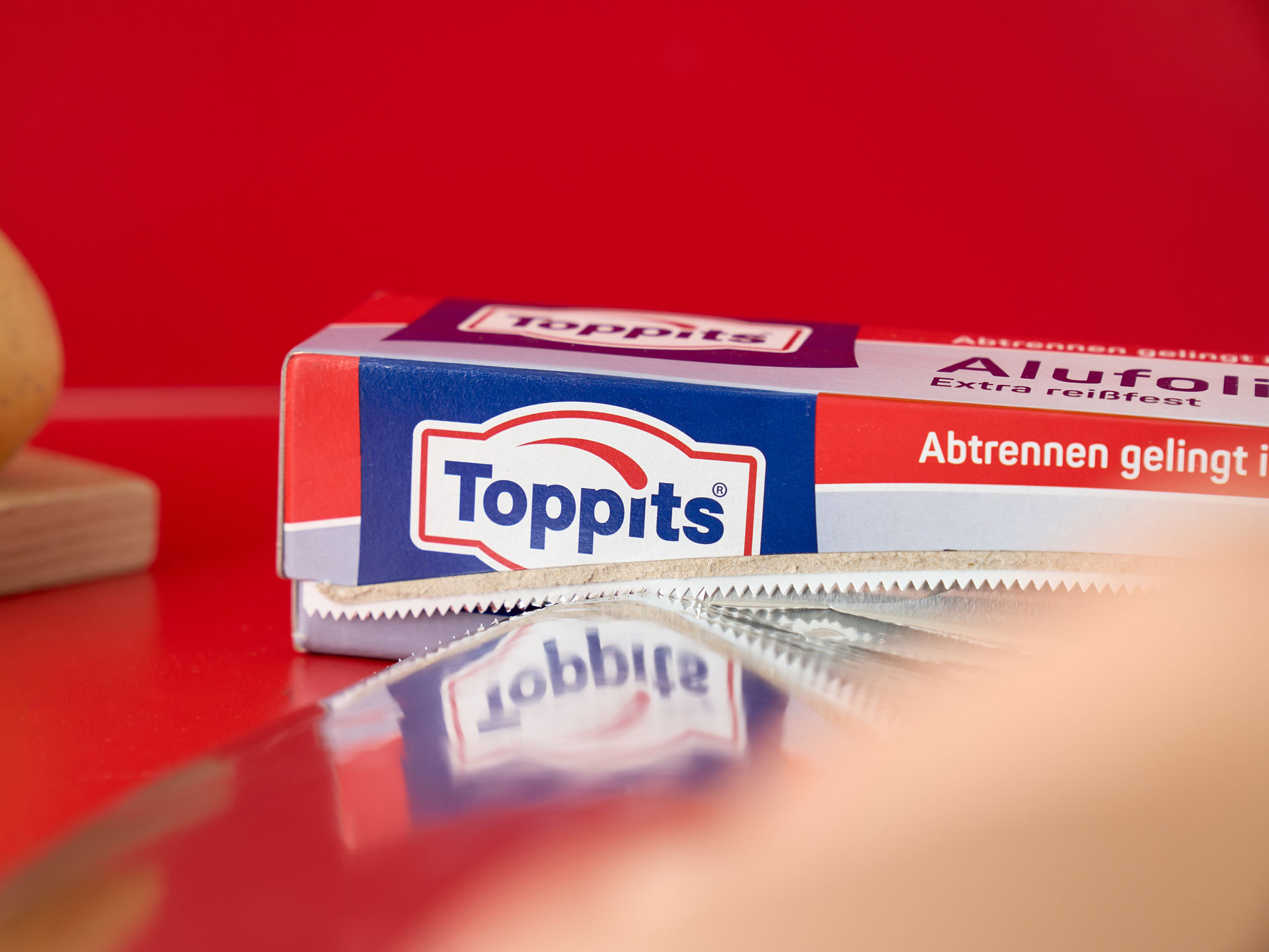 Eine Toppits Alufolien Verpackung mit herausgezogener Alufolie auf rotem Untergrund, daneben eine Kartoffel auf einem Holzbrett