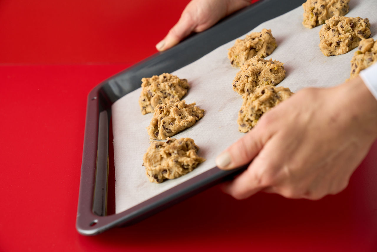 Hände halten ein Backblech mit Keksteig auf Toppits weißem Backpapier über rotem Hintergrund.