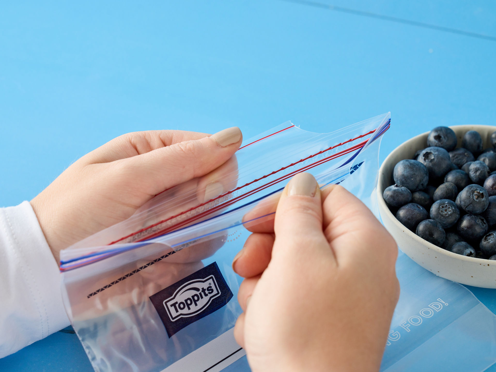 Zwei Hände öffnen einen leeren Toppits SafeLoc Gefrierbeutel, daneben steht eine Schale mit frischen Heidelbeeren auf einem blauen Untergrund