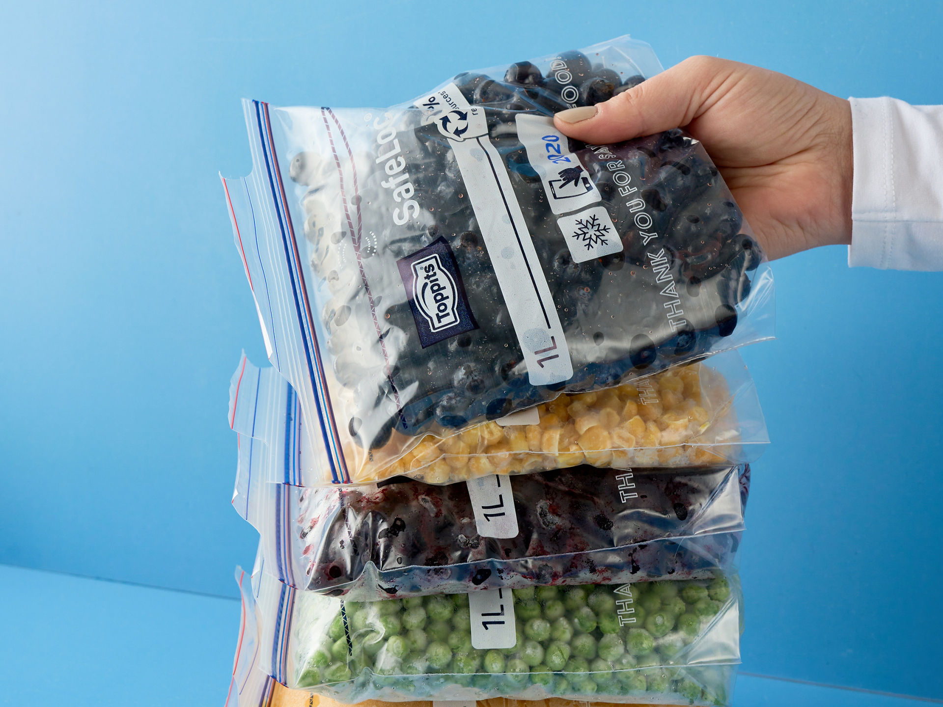Hand legt einen Toppits SafeLoc Gefrierbeutel mit Beeren, auf einen Stapel von anderen Toppits SafeLoc Gefrierbeutel die mit verschiedenen Lebensmitteln gefüllt sind, die auf blauem Hintergrund liegen