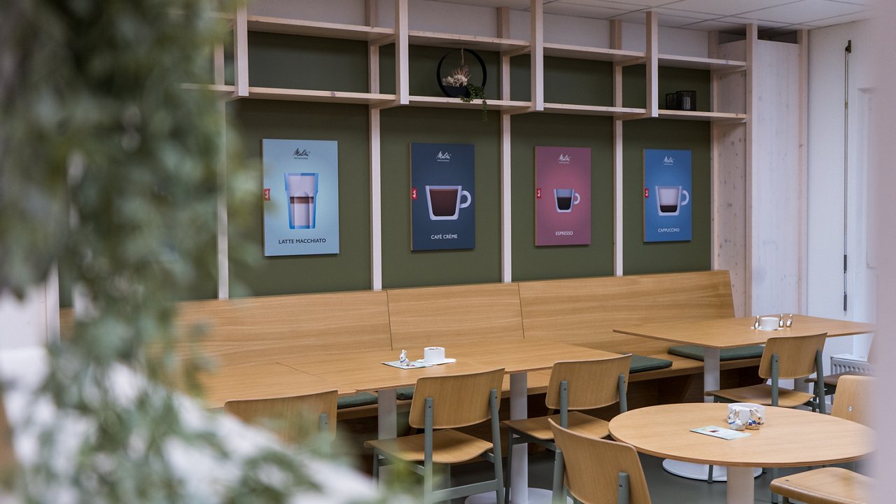 Lange Holzbank vor grüner Wand mit Kaffeegrafiken. Davor Tische und Stühle aus Holz.