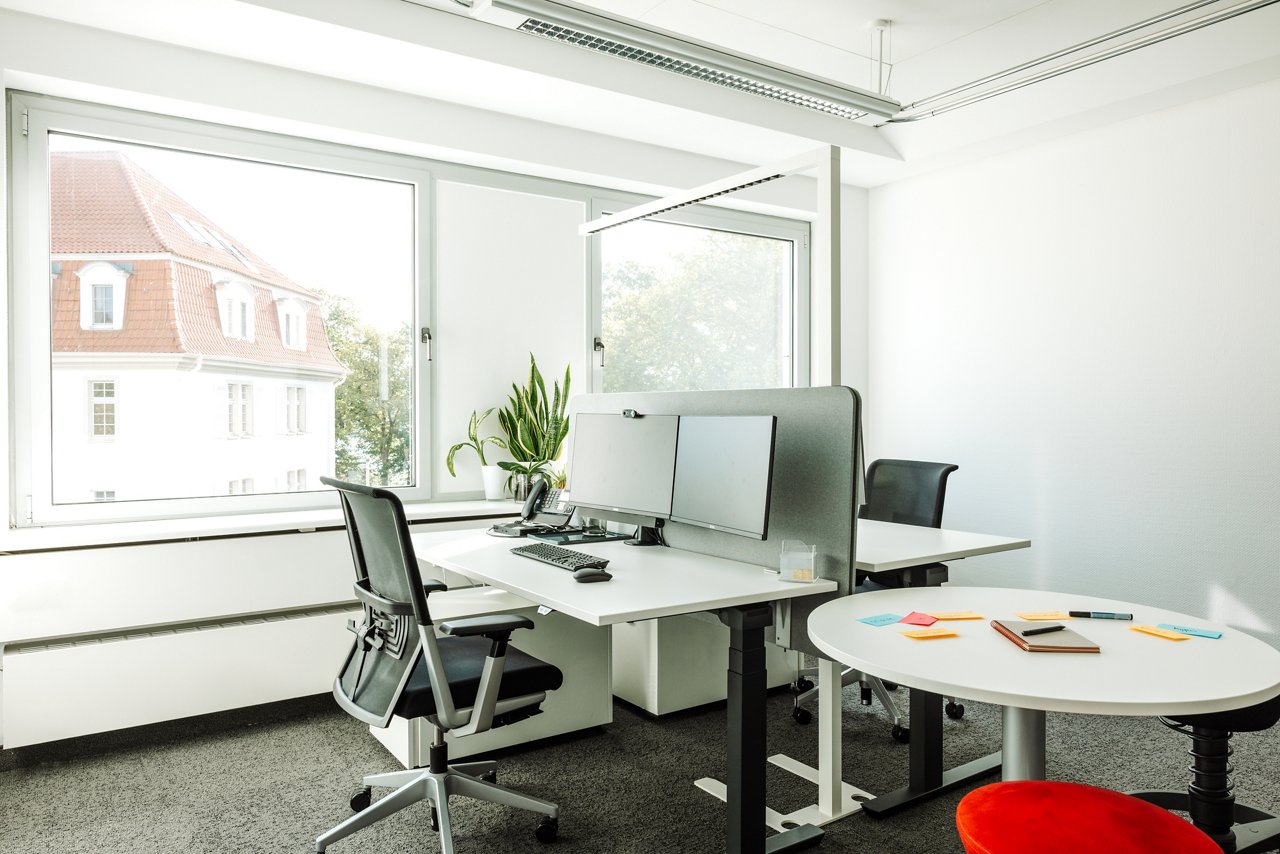An office with two workstations, with screens, desk chairs and a small round meeting table with red stools.