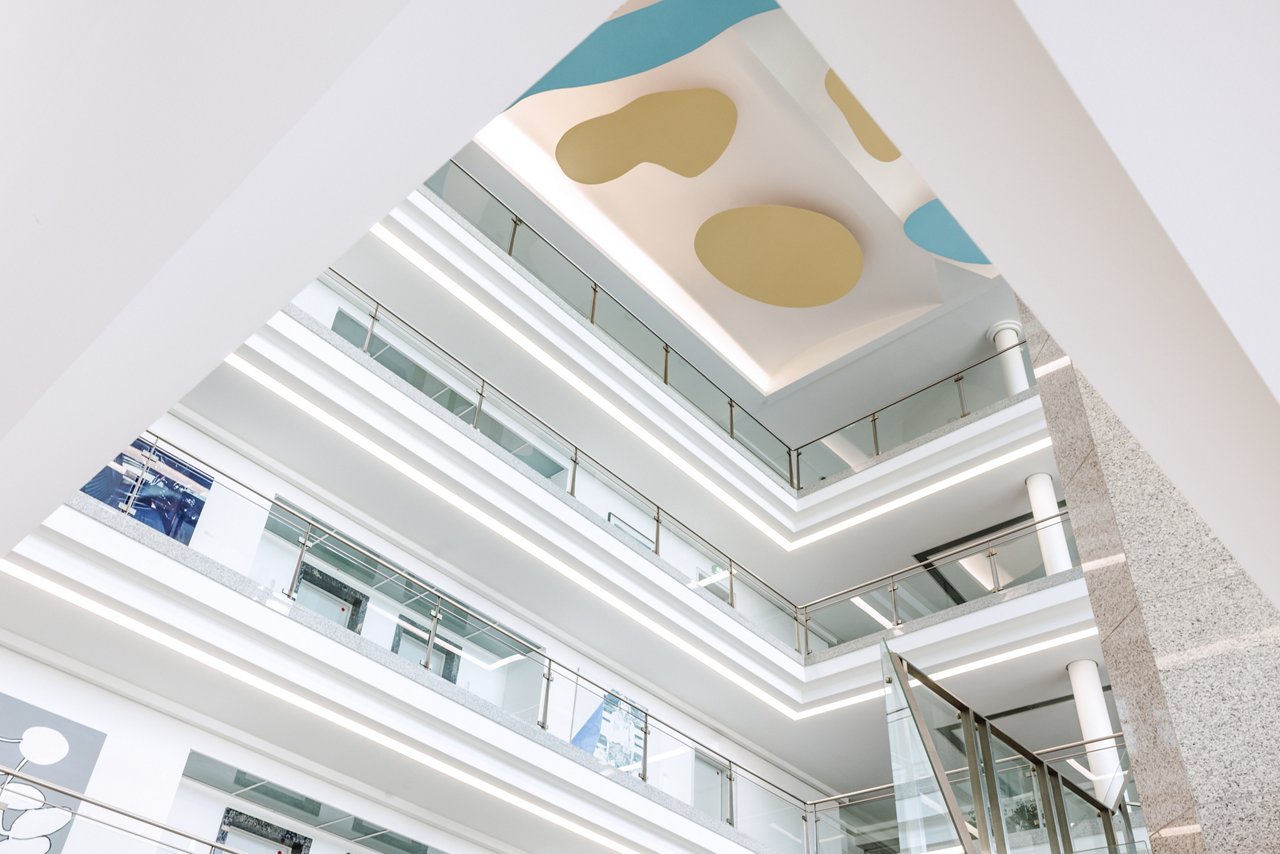 Ein helles Foyer mit vier Etagen. Der Blick geht nach oben bis zur Decke, an der gelbe und hellblaue Skulpturen hängen, die Wolken ähneln.