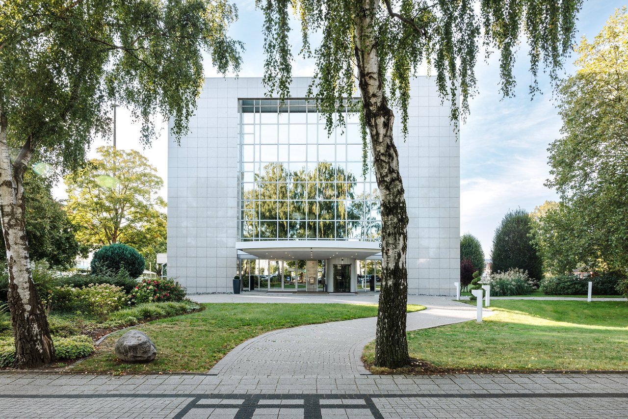 Frontal view of a building with mirrored façade framed by green landscape and a paved path winding towards the building