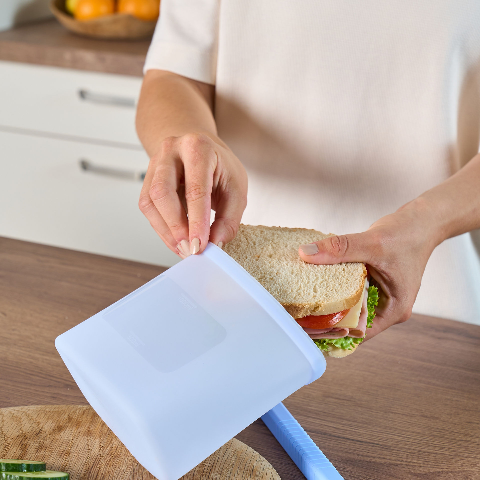 Eine Person hält ein Sandwich und öffnet einen wiederverwendbaren blauen Toppits Silikonbeutel in einer Küche, daneben ein Schneidebrett mit Gurken- und Tomatenscheiben