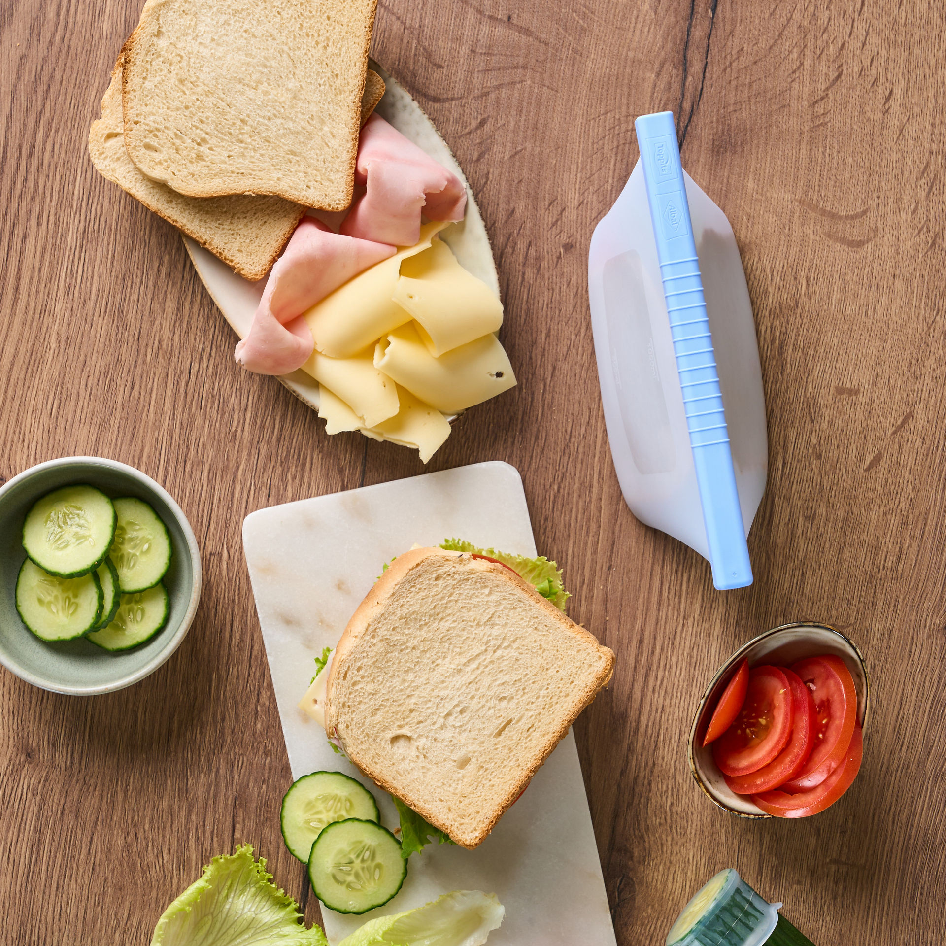 Zutaten für ein Sandwich, darunter Toastbrot, Käse, Schinken, Salat, Tomaten und Gurken, liegen auf einem Holztisch zusammen mit  wiederverwendbaren  Toppits Silikonbeuteln