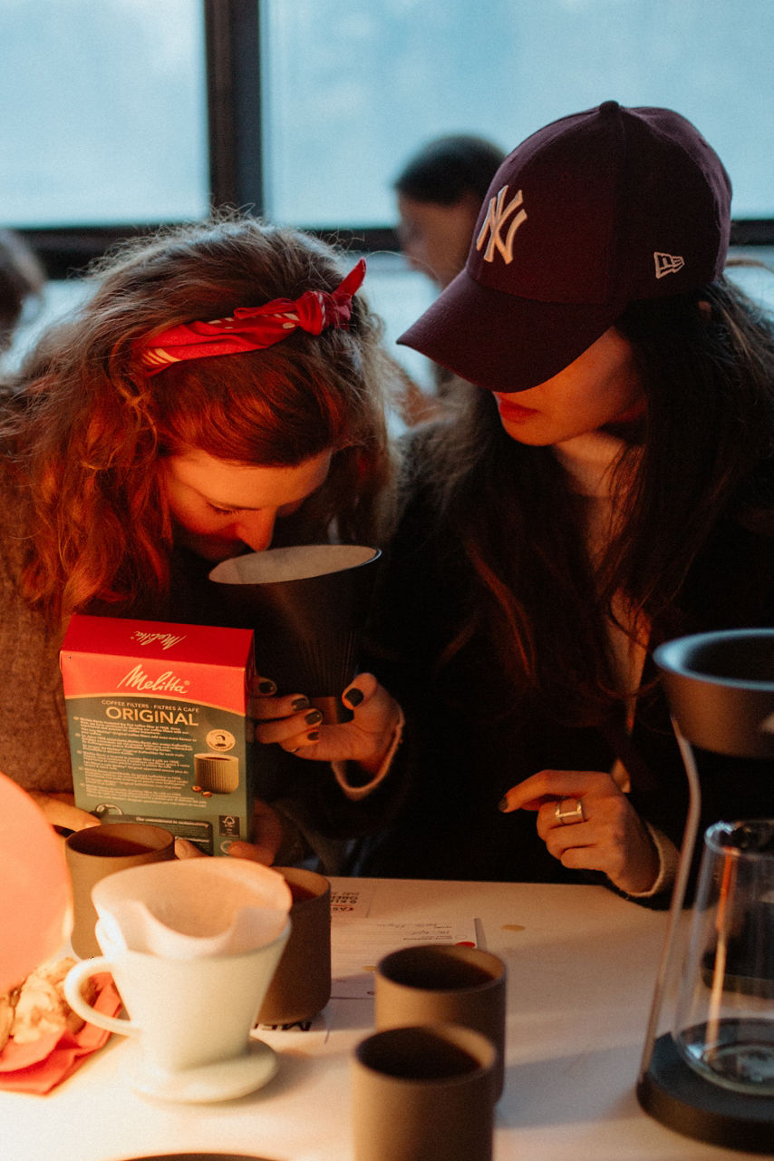 Eine Person mit rotem Haarband und Basecap beugt sich über eine Kaffeetasse und eine Kaffeemaschine; im Vordergrund eine weiße Tasse. 