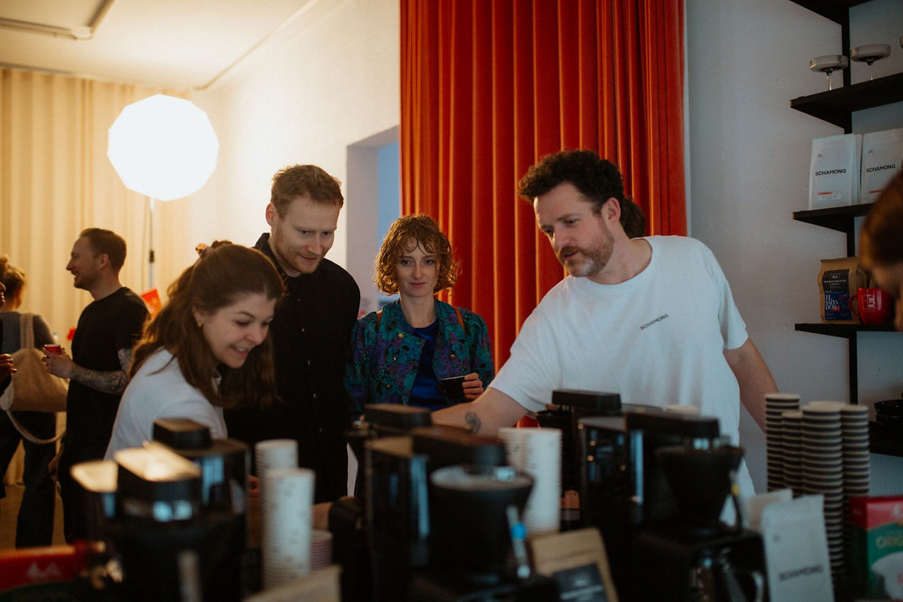 Ein Mann und eine Frau stehen nebeneinander vor einer Kaffeemaschine in einem Raum mit rotem Vorhang und hellen Kugellampen. 