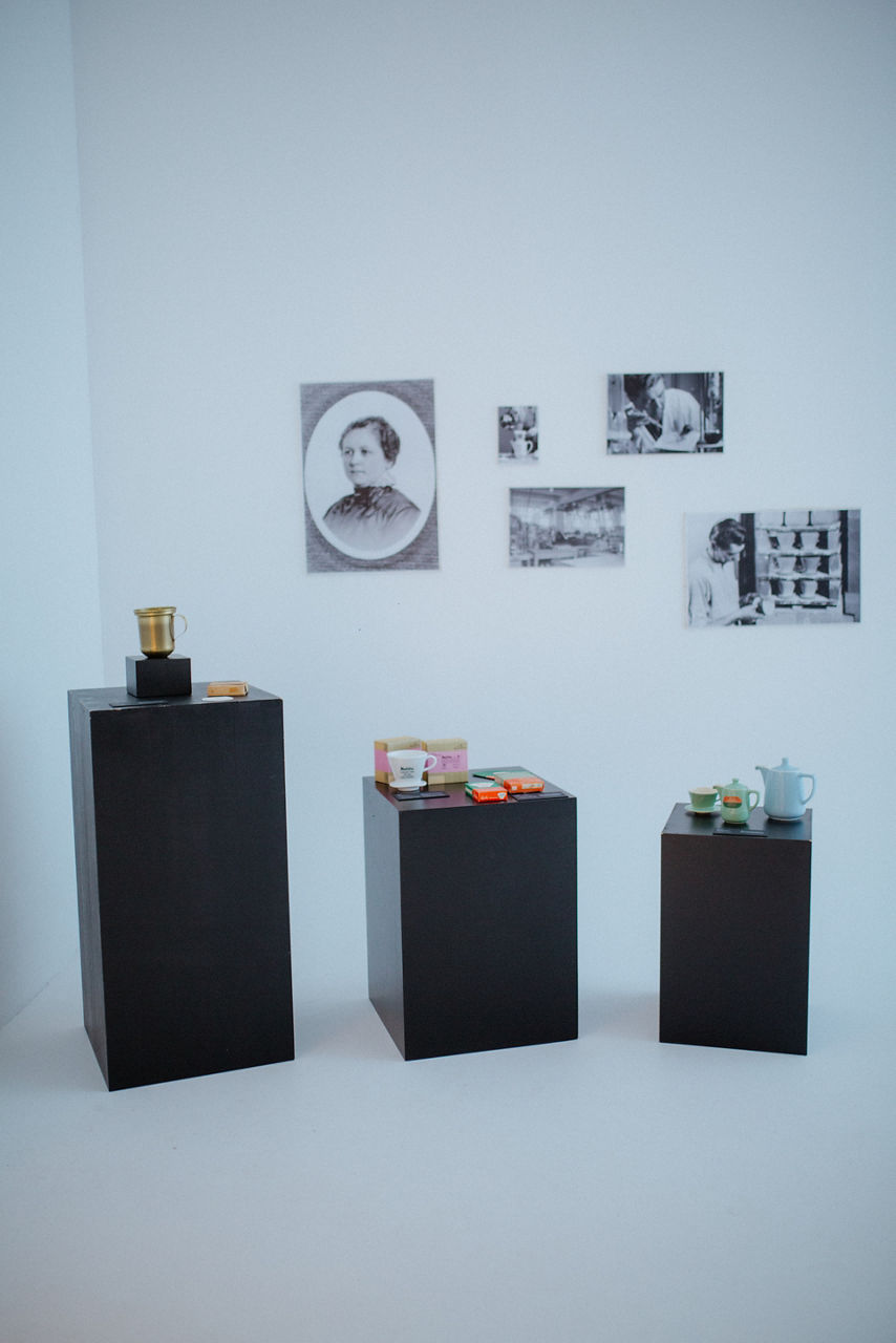 Zwei schwarze Kaffeemaschinen stehen auf dunklen Sockeln vor einer hellen Wand; im Hintergrund sind gerahmte Bilder zu sehen. 