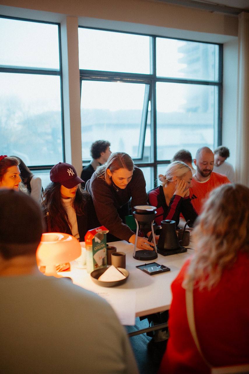 Gruppe von Personen sitzt und steht an Tischen in einem hellen Raum mit großen Fenstern; auf den Tischen stehen Kaffeemaschinen und Utensilien. 