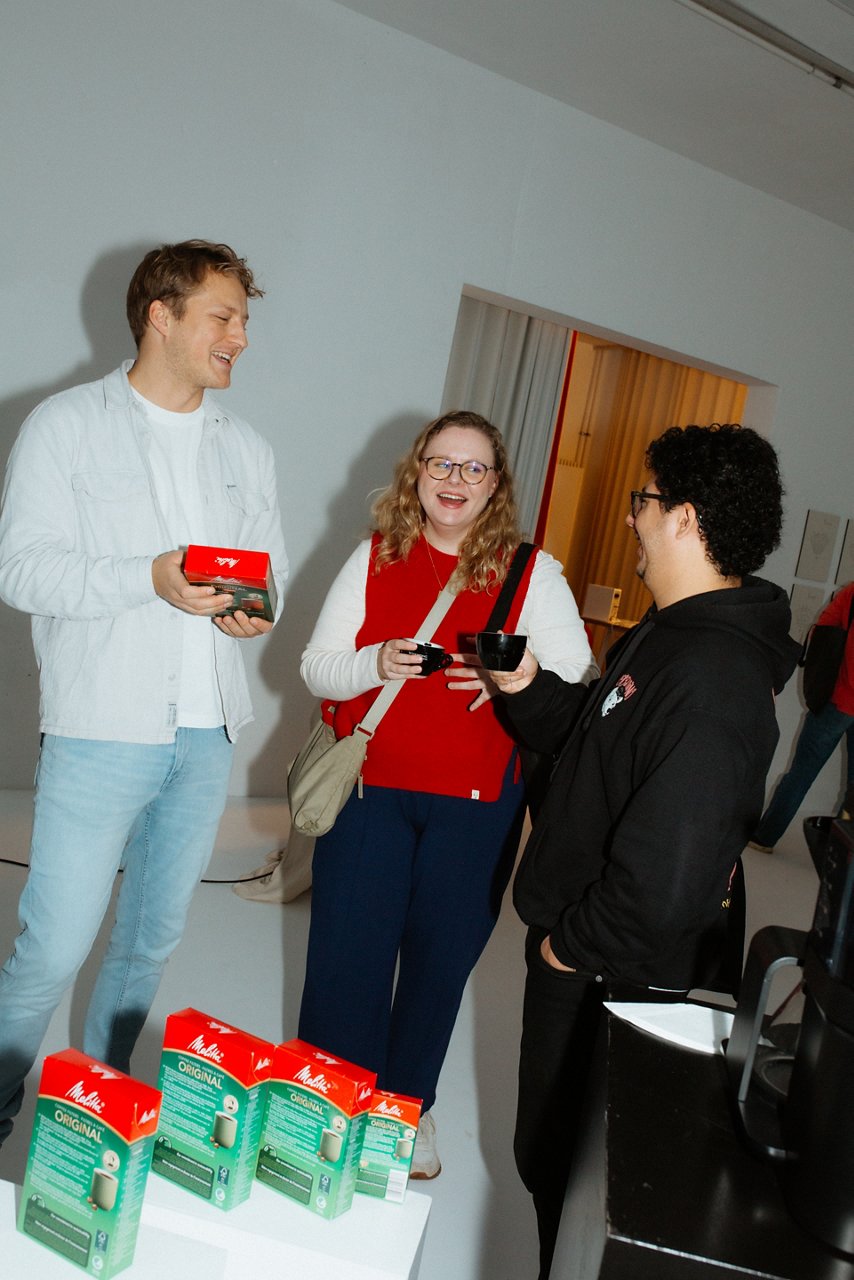 Drei Personen stehen in einem Raum und unterhalten sich; eine Person hält eine rote Tasse, im Vordergrund rote Produktverpackungen. 