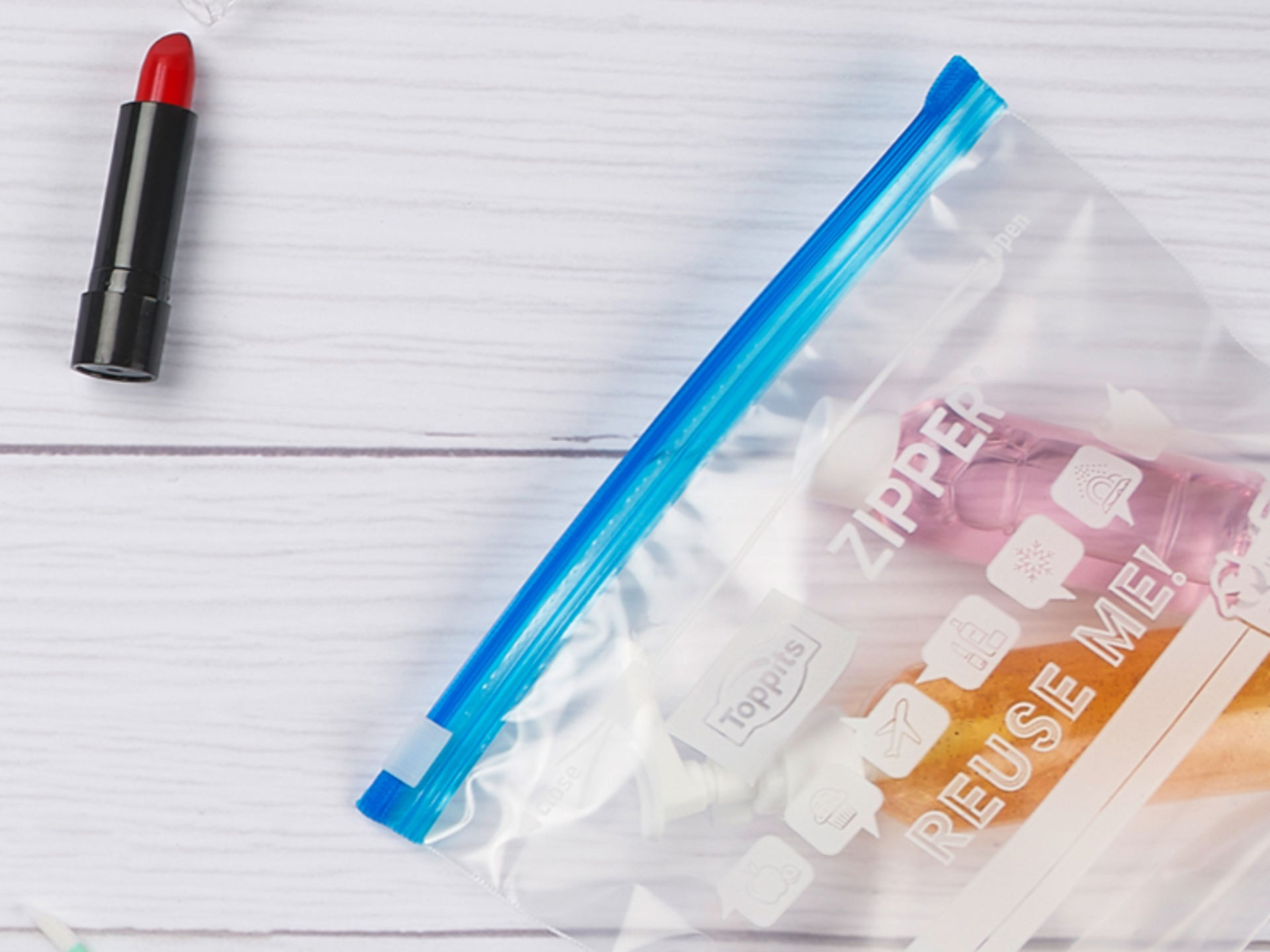 Transparenter Zipper-Beutel von Toppits mit blauem Verschluss, darin befinden sich Reisegrößen von Kosmetikprodukten, daneben liegen ein Lippenstift, Wattestäbchen und ein Puder auf weißem Untergrund