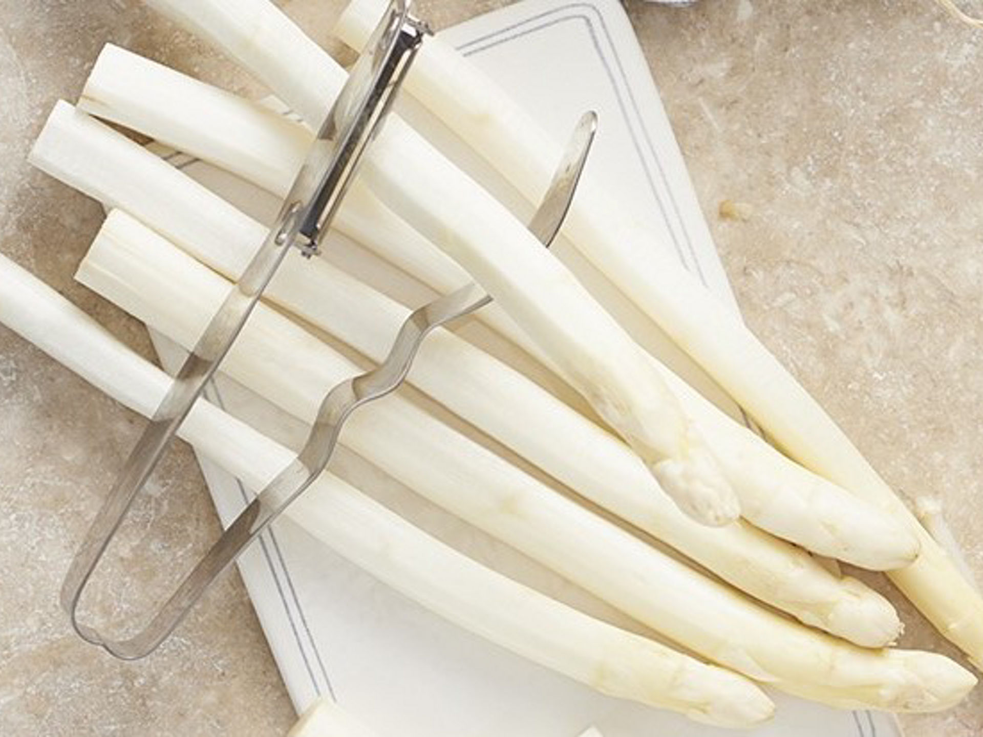 Geschälter weißer Spargel liegt auf einem Schneidebrett, daneben ein Spargelschäler auf einer hellen Arbeitsfläche