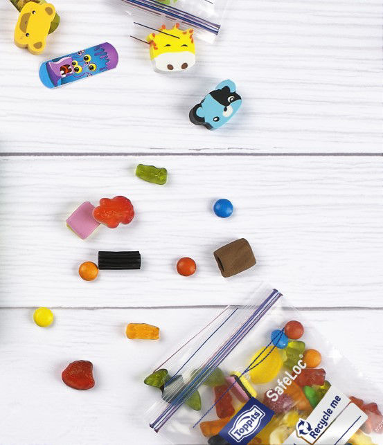 Auf einer weißen Holzoberfläche liegen bunte Radiergummis in Tierform und verschiedene Süßigkeiten. Ein Safeloc Mini von Toppits ist mit Süßigkeiten gefüllt 