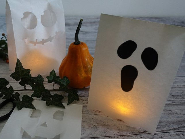 Zwei leuchtende Halloween-Lichttüten aus Toppits Butterbrotpapier mit ausgeschnittenen Gesichtern stehen auf einem Holztisch. Daneben liegen ein Zierkürbis, eine Efeuranke und ein weiteres ausgeschnittenes Papier. 