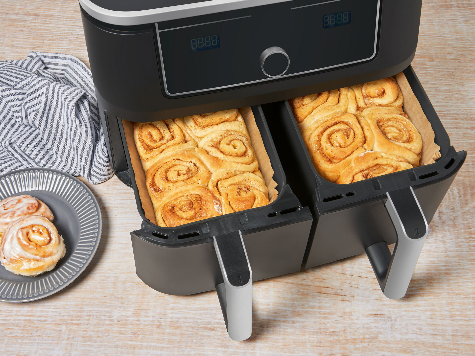 Zwei Fächer einer Heißluftfritteuse mit Zimtschnecken auf Backpapier; daneben ein Teller mit zwei Zimtschnecken. 