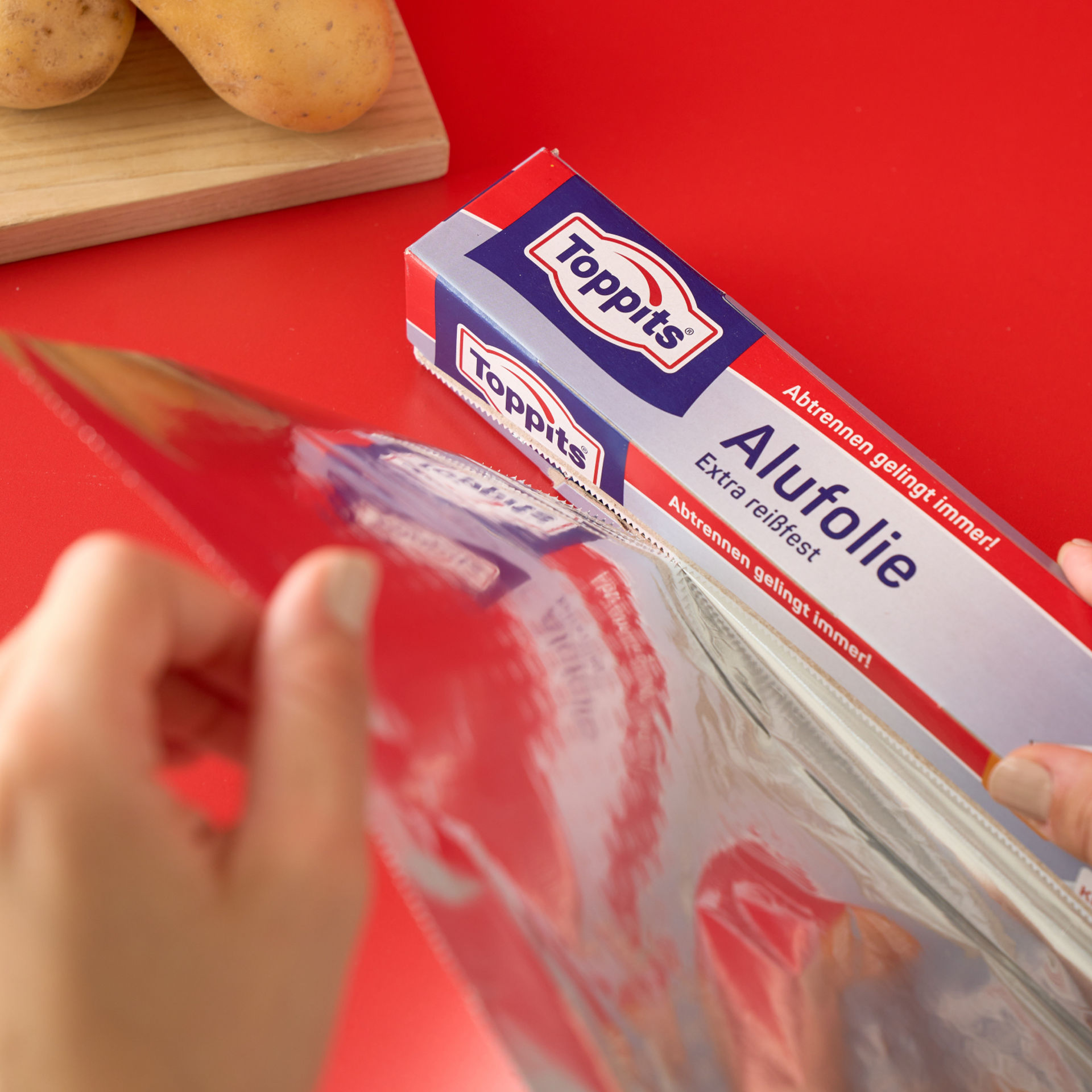 Hände ziehen Toppits Alufolie aus der Verpackung auf rotem Hintergrund.