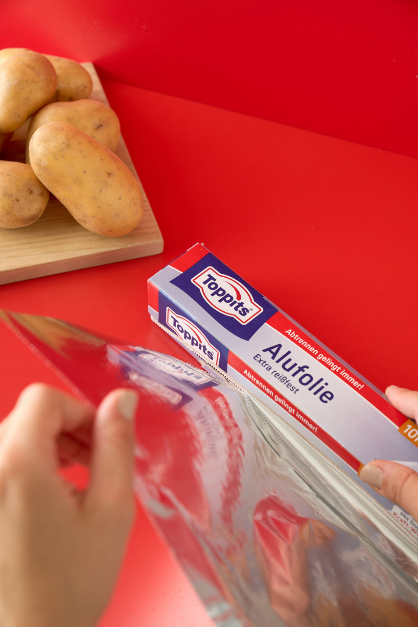 Hände ziehen Toppits Alufolie aus der Verpackung auf rotem Hintergrund.