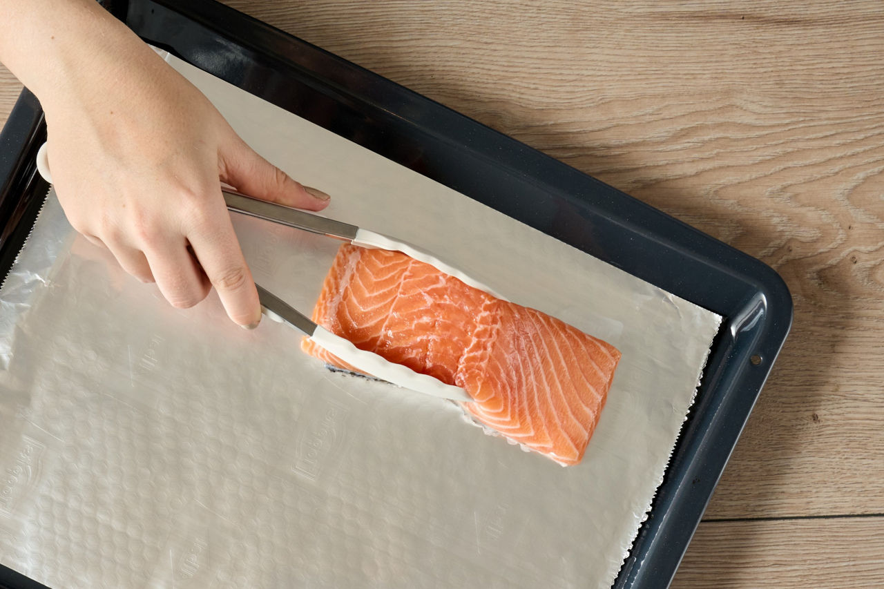 Eine Hand legt mit einer Zange ein Stück rohen Lachs auf ein Backblech, das mit Toppits Alufolie ausgelegt ist