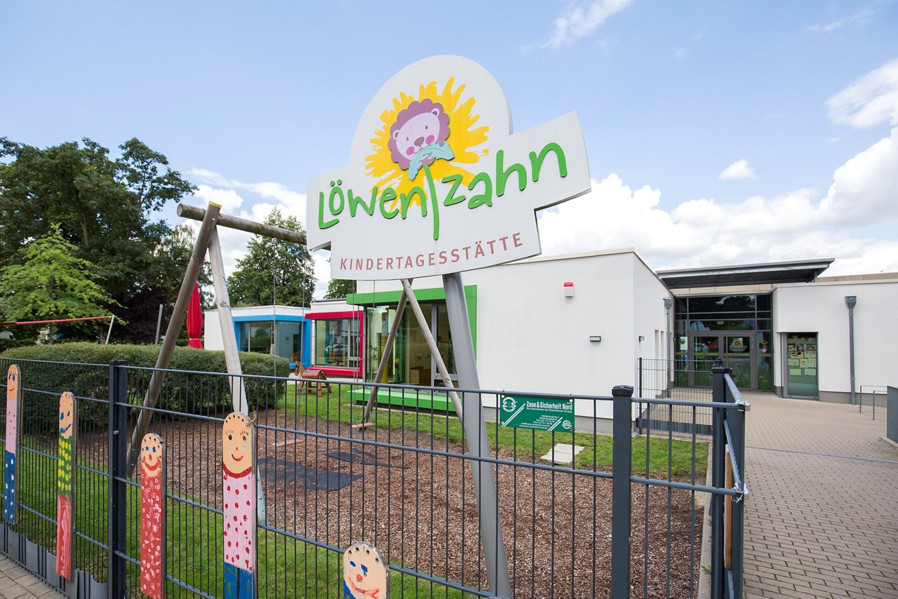KiTa Dandelion, white building from the outside with a swing and a tooth in front of it. There is a sign on the fence with the name and logo of the daycare center. 