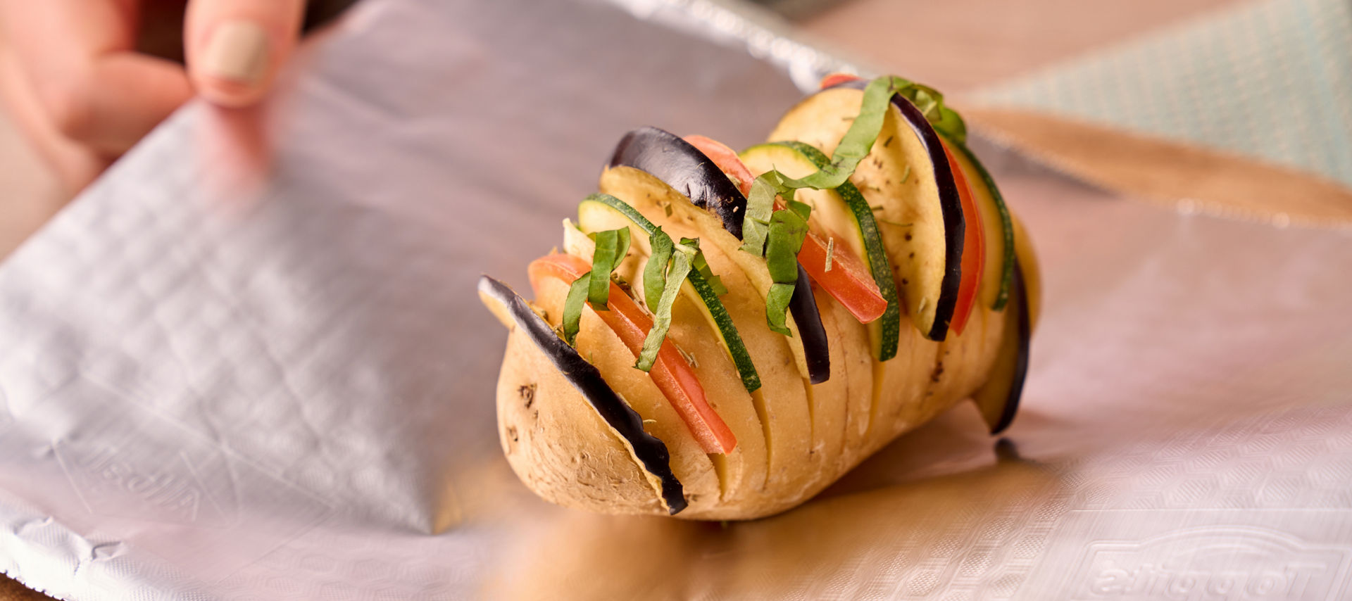 Een in plakjes gesneden aardappel, gevuld met verschillende groenten, ligt op een vel aluminiumfolie en is klaar om te worden ingepakt.