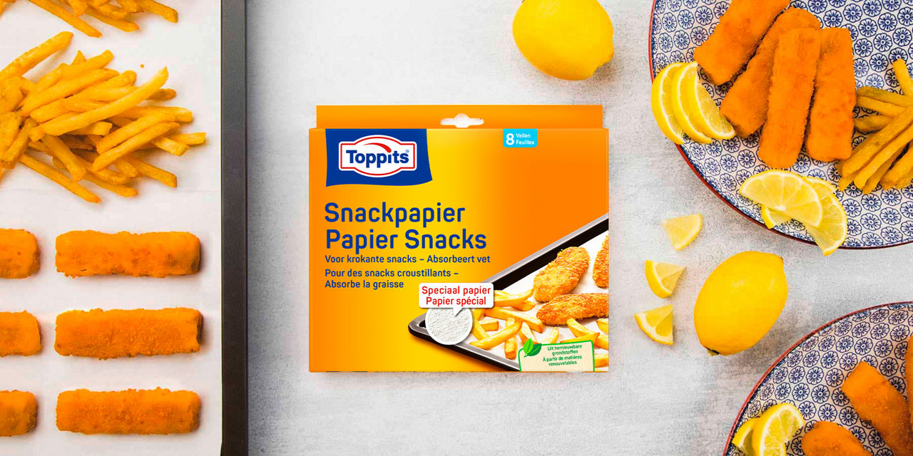 Een verpakking Toppits snackpapier ligt centraal op tafel, omringd door een bakplaat met frietjes en vissticks, een bord met citroenpartjes en gefrituurde snacks, en losse citroenen.
