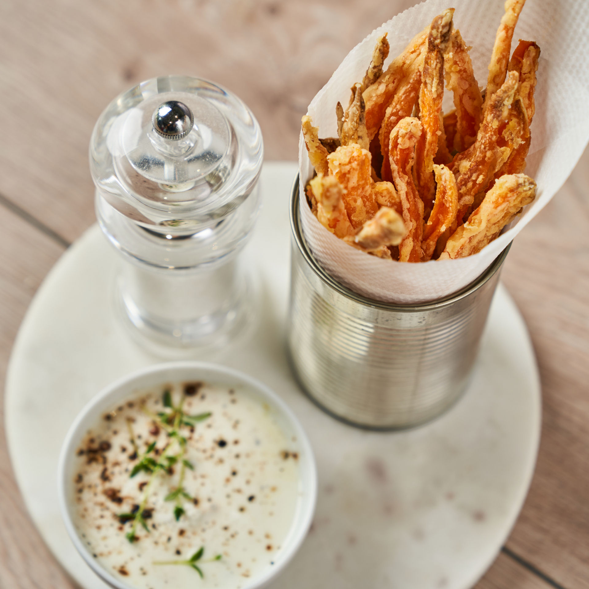 Een blik gevuld met wortelfrietjes op snackpapier staat op een wit bord naast een potje romige saus met verse kruiden en een pepermolen.