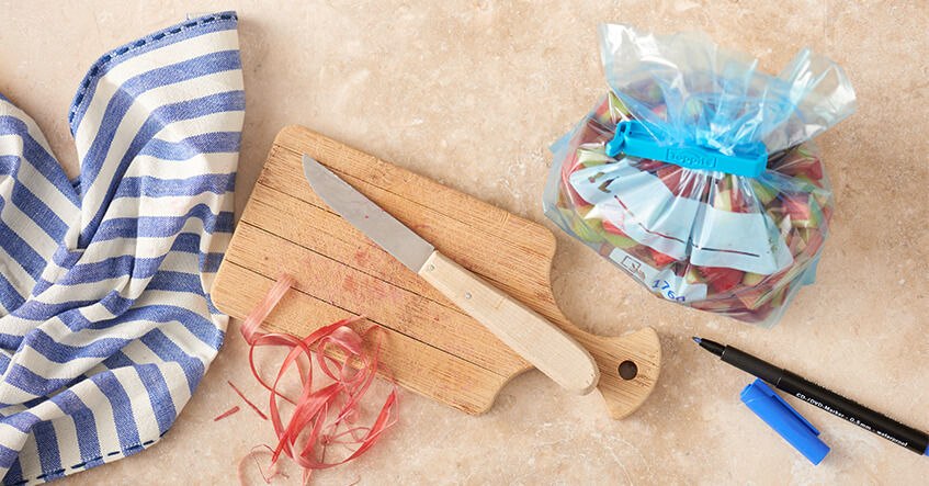 Een houten snijplank met een mes en groenteschillen ligt naast een diepvrieszak gevuld met stukjes groente, afgesloten met een blauwe klem, en daarnaast liggen een stift en een blauwe dop op het aanrecht.