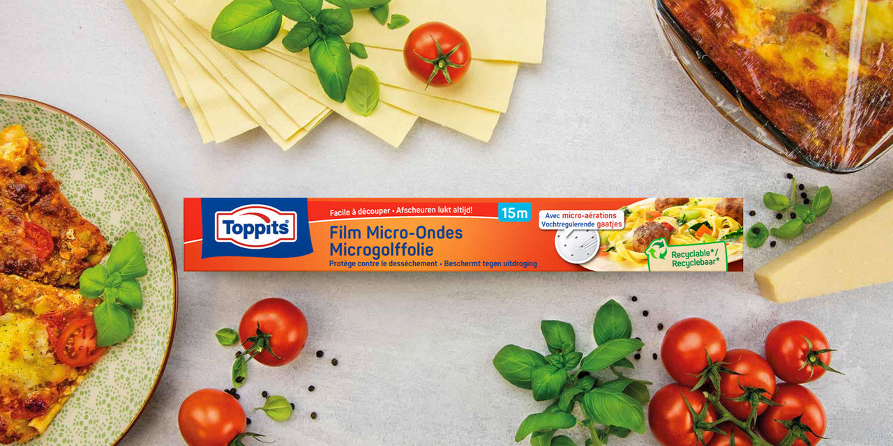 Een verpakking Toppits Microgolffolie ligt centraal op tafel, omringd door verse lasagnevellen, tomaten, basilicum, een stuk kaas en een schaal lasagne die is afgedekt met folie.